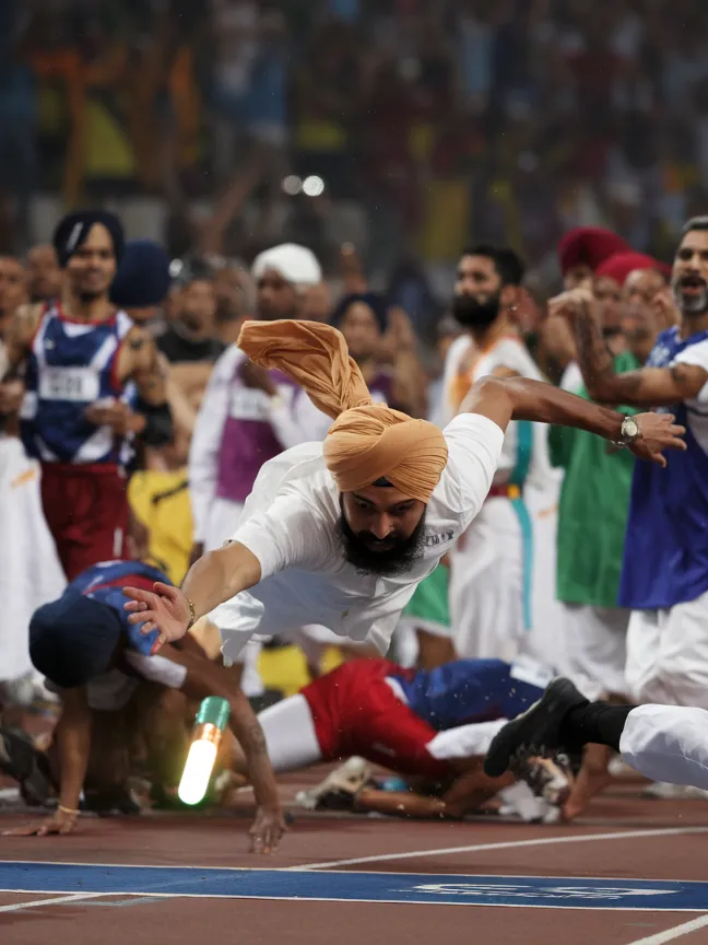 Athletes in religious garb participate in a chaotic relay race, fumbling a glowing baton shaped like clasped hands. The crowd goes wild as a Sikh runner dives across the finish line, turban unraveling in slow motion.