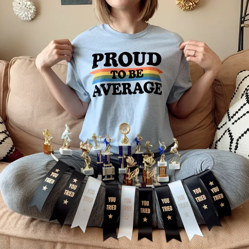  A person wearing a "Proud to be Average" t-shirt, sitting contentedly on a beige couch, surrounded by a collection of participation trophies and "You Tried" ribbons