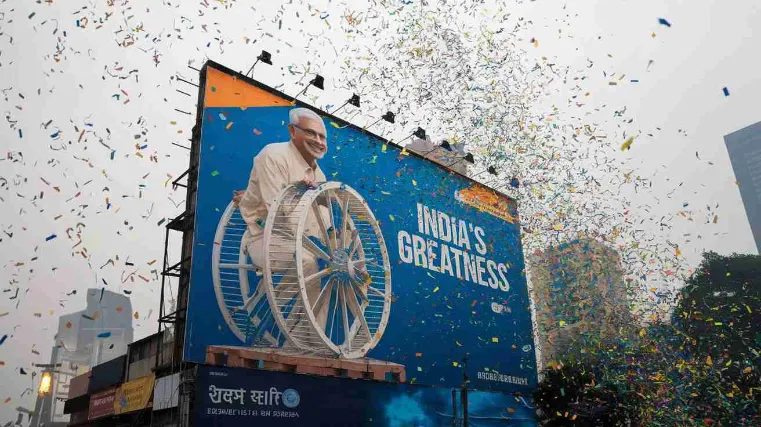 A massive billboard showing a grinning politician riding a hamster wheel labeled "India's Greatness," with confetti raining down endlessly