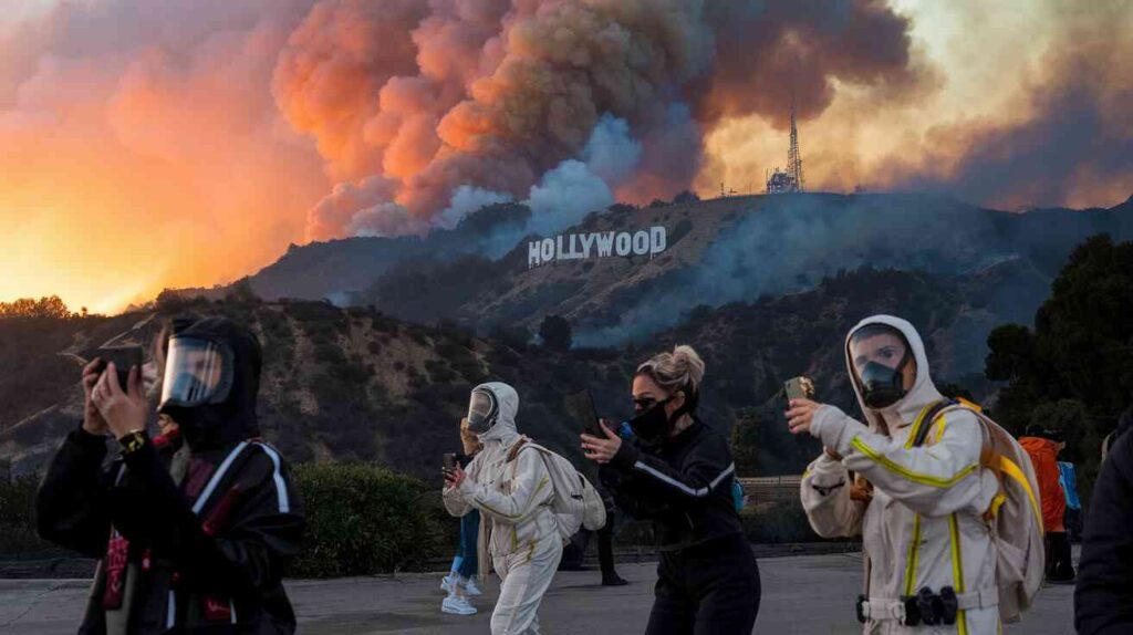 A dramatic sunset over Hollywood Hills with smoke billowing around the Hollywood sign, while influencers desperately try to find the perfect angle while wearing full face masks and designer fire-resistant suits